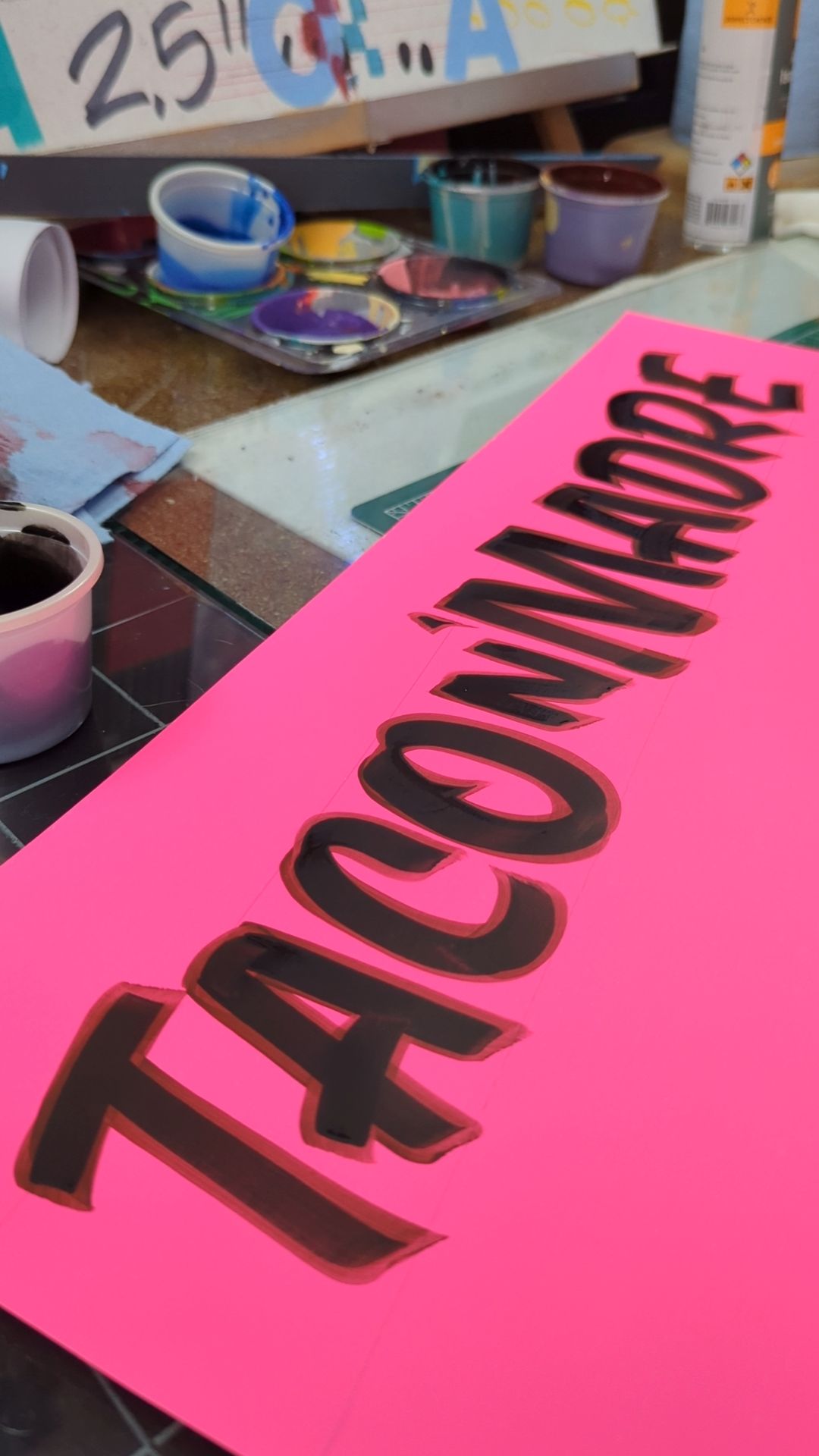 Cartulina fosforescente rosa rotulada a mano con marcador: Taco’nmadre, junto a los botes de pintura del taller